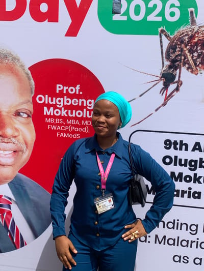 The image, rotated 90 degrees counter-clockwise, captures a smiling woman standing in front of a large event banner. She is dressed in a navy blue long-sleeved top and matching trousers, complemented by a light blue headwrap. A black shoulder bag is slung over her left shoulder, and a gold-colored ring with a butterfly design adorns her right hand, which rests on her hip. An ID badge, displaying "University of Ilorin" on its pink lanyard and a long numeric code "5078700218520749369" along with "Cerve", is clipped to her top.

The banner behind her is predominantly white with red and green accents. It features a large, detailed close-up of a mosquito, and the partially visible numbers "2026" in green. Text on the banner includes "Prof. Olugbenga Mokuolu" along with his qualifications: "MB;BS., MBA, MD, FWACP(Paed), FAMedS". Other partial texts visible are "9th A", "Olugb", "Mok", "aria", "nding I", "a: Malaria", and "as a", suggesting an event related to malaria, possibly a conference or annual gathering in Ilorin, Nigeria. A partial image of a smiling man, likely Professor Olugbenga Mokuolu, is also visible on the lower part of the banner, wearing a striped tie. No specific time of day or weather cues are evident.