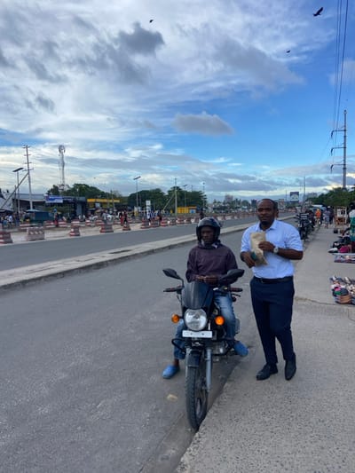 The image shows a scene in Dar es Salaam, Tanzania, taken from coordinates (latitude: -6.768, longitude: 39.226). Two men are standing on the side of a paved road, with a black motorcycle parked nearby. The man on the left is wearing a dark jacket, a helmet, and blue jeans. He is seated on the motorcycle, looking down. The man on the right is wearing a light blue shirt, dark pants, and black shoes. He is holding a brown paper bag and is looking at the camera. There are several buildings in the background, along with some traffic and street signs. The sky is blue with white clouds, and the weather appears to be sunny. There are multiple power lines in the sky and there are birds flying overhead. The location is likely a busy street in the city.