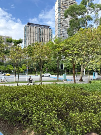In the Haizhu District, China, on a sunny day with scattered clouds, a street scene unfolds. A person rides a white electric scooter on the right side of the foreground. In the middle ground, traffic moves along a road marked with blue lanes. A yellow taxi is visible on the left, and a white car is on the right. Tall modern buildings, some with balconies and glass facades, rise in the background, interspersed with lush green trees. Charging stations for electric vehicles are present near the road. The foreground is dominated by dense, manicured green bushes and manicured grass.