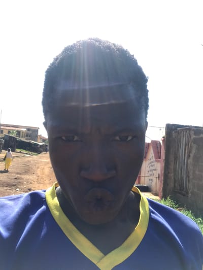 The image displays a close-up, rotated portrait of a person taking a selfie in an outdoor, brightly lit setting. The subject, who appears to be a young individual with dark skin and short, dark, curly hair, occupies the left side of the frame. They are wearing a blue shirt with a yellow trim. Their eyes are open and directed at the camera, and their lips are pursed in a pouting expression.
Strong sunlight is evident, creating significant lens flares and glare on the left side of the subject's head, indicating a clear, sunny day, likely around midday or early afternoon. The background reveals an urban or semi-urban environment with buildings and unpaved ground. To the right, behind the subject, a reddish-brown building with visible text inscribed vertically on its wall can be seen. Phrases discernible include "The Living God," "No meaning," "god background," and "SAVE LIFE."
The ground beneath the buildings appears to be dirt or gravel. In the lower right corner of the frame, a smaller figure of another person, dressed in yellow, is visible walking in the distance. The scene collectively suggests a location consistent with Mandate Estate, Ilorin, Nigeria.