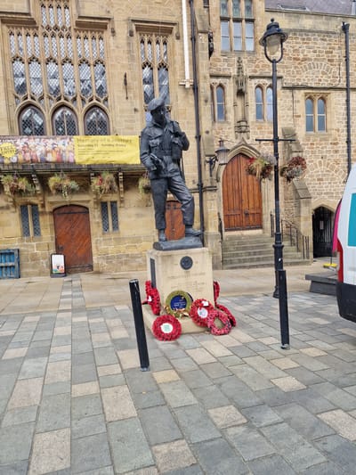 The image shows a war memorial in the city of Durham, United Kingdom. The statue of a soldier stands in front of Durham Town Hall. A banner to the left of the statue advertises an event called "Memory of a Journey" at the town hall from Saturday 23 to Sunday 31 May. Wreaths of poppies are placed on the memorial. The ground is paved with rectangular gray and tan stones. A lamp post is to the left of the memorial. A partial view of a vehicle is visible at the top right corner of the image.
