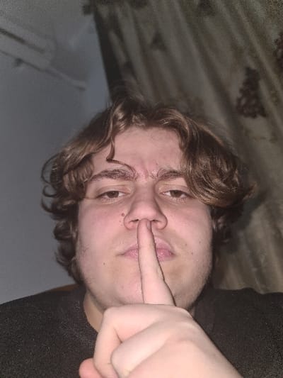 A person with wavy brown hair and a light complexion is holding a finger up to their nose. The person is wearing a dark shirt. The background includes a textured wall or fabric behind their head and a plain wall to the left. The image is slightly rotated, giving the impression of looking up at the person. Their eyes are visible, and they appear to be looking towards the camera. The lighting is somewhat dim, casting shadows across the face and background.