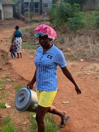 A person with a pink hair bonnet, blue striped shirt, and yellow shorts stands outdoors. They hold a gray trash can and wear flip-flops. Two people, an adult and child, walk in the background on a dirt path. Grass and trees surround the scene. Buildings are visible in the distance.