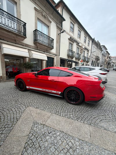 The image depicts a red Ford Mustang 5.0 coupe parked on a cobblestone street in Vila Real, Portugal. The vehicle is angled slightly towards the right, occupying the left side of the street, and features black multi-spoke wheels with red trim and a white "MUSTANG 5.0" decal along its lower side. Behind the Mustang, a white SUV is parked, followed by additional vehicles further down the street. The street surface consists of traditional Portuguese cobblestone, laid in a fanning pattern, with sections of lighter, larger granite slabs in the foreground.
The left side of the street is lined with multi-story buildings of traditional Portuguese architecture. The building immediately to the left of the Mustang features light-colored facades, ornate stone window surrounds, and black wrought iron balconies. Its ground floor houses a commercial establishment with large glass windows and two extended beige awnings. Reflections in the windows indicate a business named "Central" and an interior display featuring another red vehicle. Adjacent to this, another building with similar architectural characteristics displays a sign for "CALDEIRAS" and includes a visible ATM on its facade. The sky overhead is overcast. No individuals are visible in the scene.