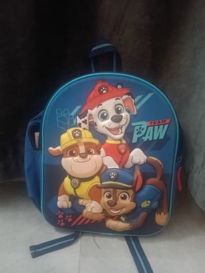A blue Paw Patrol backpack is shown in a close-up shot. The backpack features characters from the show, including Marshall, Rubble, and Chase, depicted in a cartoon style. The background is dark and out of focus, with a hint of a gray textured surface to the right. The backpack is placed on a floor with a tiled pattern, and a strap of the backpack is visible extending towards the bottom of the frame. There are no people or specific city landmarks visible to indicate a location in Chișinău, Moldova.