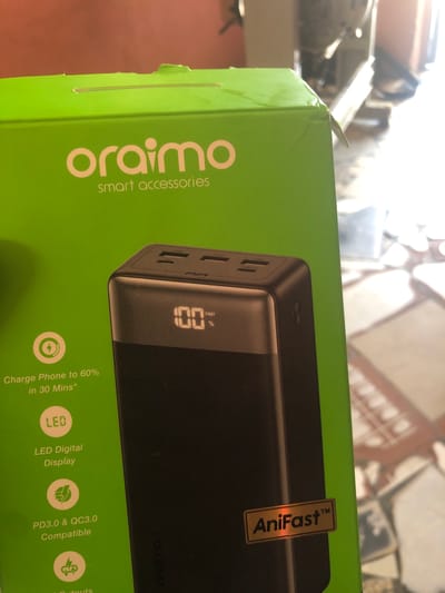 This image displays an "oraimo" brand power bank box, set against a blurry indoor background, possibly in Ado, Nigeria. The box is predominantly green, with the "oraimo smart accessories" logo prominently featured on its left side.

The main subject is a large, high-resolution image of a black power bank printed on the box. This power bank features a digital display showing "100 FAST %", indicating a full charge and fast charging capability. Multiple charging ports are visible on the side of the power bank in the depiction. A golden sticker with "AniFast™" is placed on the power bank's image on the box, and the word "AniFast" is also visible on the power bank's surface itself.

Several features of the power bank are listed in white text on the green background:
*   "Charge Phone to 60% in 30 Mins*"
*   "LED Digital Display" (with an icon showing "LED")
*   "PD3.0 & QC3.0 Compatible" (with a lightning bolt icon)
*   The word "outputs" is partially visible at the bottom right.

The top left corner of the box shows minor creasing or damage, suggesting it might have been handled or stored. The background is out of focus, revealing hints of light-colored objects and possibly a wall or patterned surface, illuminated by what appears to be natural daylight.
