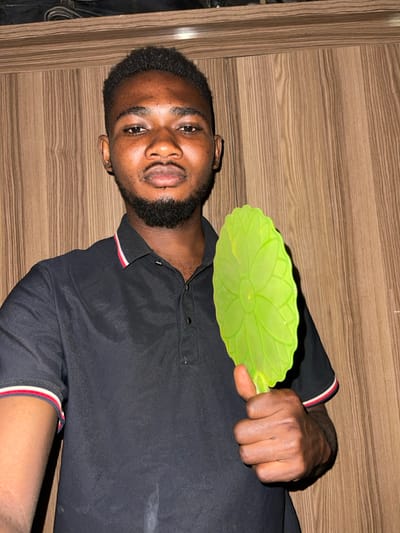 A dark-skinned man with a beard poses in front of a wooden-looking background. He wears a black polo shirt with white and red accents on the sleeves and collar. He holds a green, lotus-shaped silicone lid in his right hand, displaying it towards the camera. The image is rotated, positioning the man on his side.