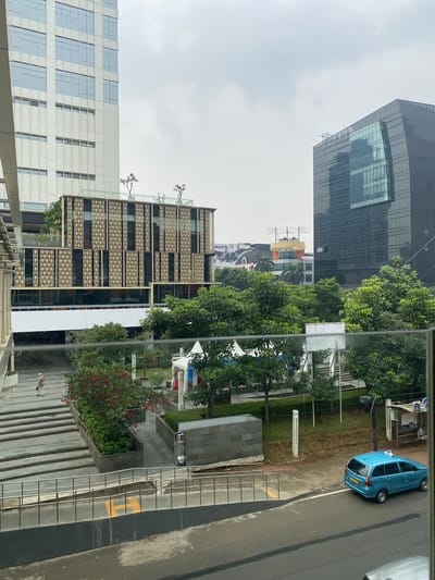 The image captures a street-level view of a modern urban environment in Central Jakarta, Indonesia, with a focus on architectural elements, greenery, and traffic. In the foreground, a blue taxi with a light blue roof sign is parked on the right side of a wet asphalt road. Adjacent to the road is a sidewalk with metal railings, behind which is a landscaped area with manicured bushes and trees.
Dominating the background are several contemporary buildings. On the left, a tall, white skyscraper with a grid of windows stretches upwards. To its right, a mid-rise building features a distinctive gold-colored geometric facade. Further to the right, a dark, curved glass skyscraper reflects the overcast sky. A few white tents are visible in a park-like area between the buildings, suggesting a possible event or outdoor market. A lone person in a light-colored outfit walks up a set of wide stairs on the left side of the frame, moving away from the camera. The overall impression is a busy but orderly city scene, characterized by modern architecture and urban planning, under a cloudy sky.