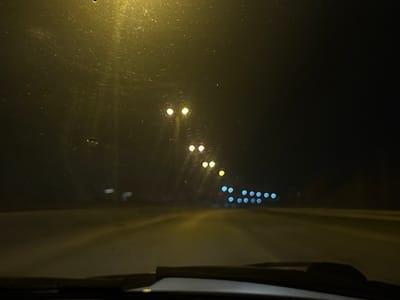 The image is a night-time view from inside a vehicle, presumably a car, driving on a road. Streetlights are visible along the side of the road, becoming more distant and blurred in the background. The windshield appears slightly dirty. The road is illuminated by the car's headlights and the streetlights. The wipers are visible at the bottom of the frame. The location is Кондратово, Russia, as stated.