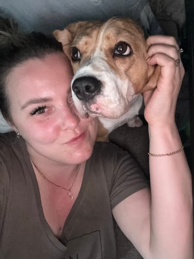 A selfie depicts a woman lying down with a dog resting its head on her forehead. The woman has fair skin, dark eyelashes, a nose piercing, and is wearing a tan shirt and gold necklace. She also has a ring on her left hand and a gold bracelet on her right wrist. The dog is light brown and white with large, expressive eyes and a black nose. The background is blurry and suggests an indoor setting, possibly a bed or couch. There is no information to suggest this image was taken in Марковский, Russia.
