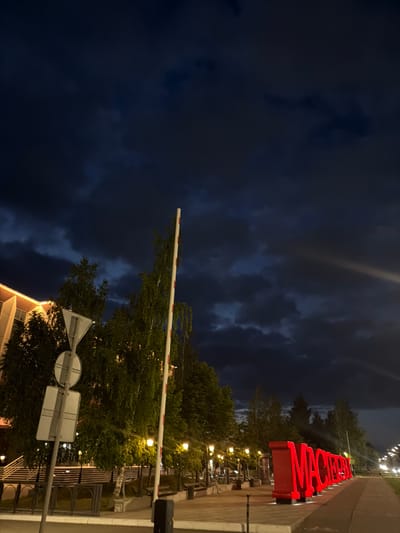 The image depicts a nighttime scene in Naberezhnye Chelny, Russia. In the foreground, large red letters spell out "МАСТЕР", illuminated from below. To the left of the letters, a white and red striped barrier stands upright, and to its left, a traffic sign with a triangular and circular plate is mounted on a pole. Behind the barrier and sign, trees with green foliage are visible, some illuminated by streetlights. A building with illuminated windows is partially visible on the far left. In the midground, a row of streetlights and benches line a paved walkway. The background shows a road stretching into the distance with more streetlights and trees. The sky above is dark with dramatic, cloudy formations. The overall impression is that of an urban public space at dusk or night, with a prominent illuminated art installation.