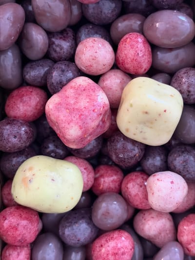 The image is a close-up, overhead shot of a pile of various-colored candies. The candies are small, rounded, and have a slightly irregular shape, suggesting they are chocolate-covered or coated in a similar confection. Colors range from light pink, deep pink, and reddish-purple to a pale yellow. Some of the candies have speckles of darker color within them, possibly indicating fruit pieces or a textured coating. The candies are densely packed together, filling the frame. No specific location in Holon, Israel is identifiable from the image itself; it is a generic depiction of candy.