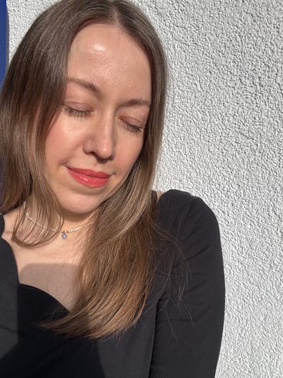 A woman with closed eyes is lying down, facing slightly towards the left. She has light brown hair and fair skin. She is wearing a black long-sleeved shirt and a delicate silver necklace with a small blue pendant. The background is a textured white surface, likely a wall. The location, Kudrovo, Russia, cannot be independently verified from the image.