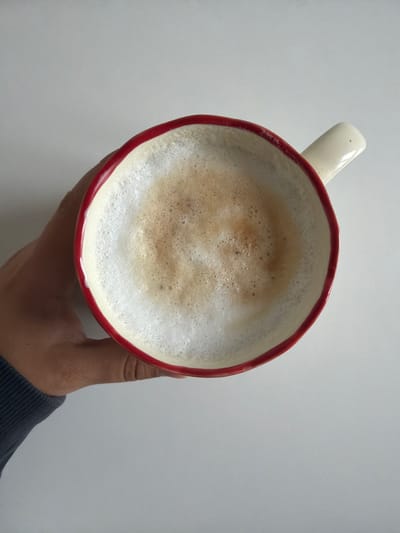 A top-down, medium shot shows a hand holding a red-rimmed white mug filled with foamy coffee. The coffee has a light brown crema. The mug has a white handle with a small red dot on it. The hand is holding the mug from the left side and is wearing a dark grey long-sleeved shirt. The background is a plain, light grey surface. The image appears to be taken indoors on a flat surface. The location is in the city of Фролы, Russia.