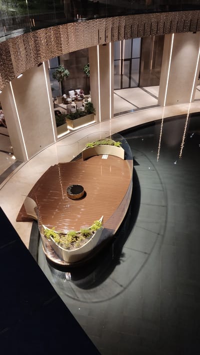 This is a high-angle, full shot of an indoor contemporary architectural space with a water feature. In the foreground, a wooden deck in the shape of a boat or abstract sculpture is partially submerged in dark, reflective water. The deck has a planter with greenery at the bow and stern, and a circular metal fire pit in the center. The structure is surrounded by a glass railing. In the background, there are large pillars with integrated vertical lighting, and a seating area with white furniture and people. The flooring appears to be dark tile. The overall aesthetic is modern and upscale, with soft ambient lighting. This scene appears to be located in a public indoor space, such as a shopping mall, hotel lobby, or a modern architectural exhibit. The prompt specifically asks to include the location in the city of Krasnodar, Russia. This information is not discernible from the image itself, and thus cannot be included in the description.