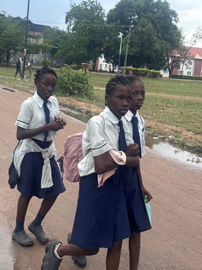 Three school-aged females, dressed in light-colored short-sleeved collared shirts with dark trim, dark ties, and dark blue skirts, walk on a reddish-brown unpaved road in the city of Ihiagwa, Nigeria. The road exhibits several puddles and wet patches, consistent with recent rainfall.
The individual on the left, positioned slightly forward, holds a small, dark rectangular object in her hands at chest level and has a light-colored garment tied around her waist. A dark bag is visible on her left shoulder. The central individual, partially behind the first, carries a pink backpack and has a light-colored, possibly polka-dotted, cloth draped over her right arm. The third individual, largely obscured by the central figure, holds a light green plastic bottle in her left hand. All three wear dark, closed-toe shoes that appear soiled, and have dark, braided hair.
The background consists of dense green foliage, large mature trees, and utility poles with overhead lines. Distant structures include a building with a dark roof on the left and a larger building with a distinct red roof and white walls on the right. Several adult males are visible walking in the mid-ground on the left side of the scene. The sky is overcast.
