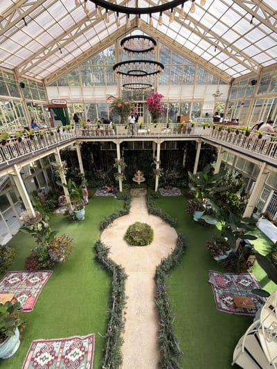 The image depicts the interior of a large, multi-level greenhouse cafe. The structure features a transparent glass roof and walls, allowing ample natural light to illuminate the space. The interior is designed with a central courtyard area covered in artificial turf.
On the ground level, there are several seating areas arranged on rugs, with plants and potted flowers surrounding them. A small fountain is visible in the center of the courtyard. A pathway made of light-colored stone winds through the artificial turf.
The upper levels consist of walkways and balconies overlooking the courtyard. These areas are lined with railings and also feature numerous potted plants. People are seen sitting at tables and conversing on these upper levels. A "Photo Booth" sign is visible on one of the upper walkways.
The overall atmosphere is lush and green, with a clear emphasis on bringing the outdoors inside. The cafe appears to be a popular gathering spot, with visitors enjoying the unique environment. The location is identified as a cafe in Baturiti, Indonesia.