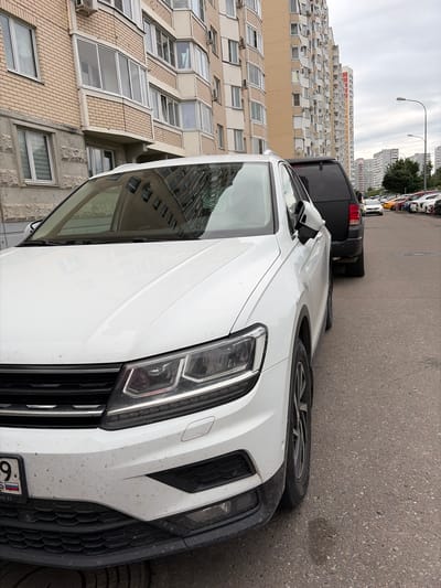 A white Volkswagen Tiguan is parked on a paved street in front of a multi-story apartment building. The car is facing towards the right of the frame, and its front bumper, headlight, and part of the hood are visible. The car appears dirty. Behind the Tiguan, a black SUV is also parked. In the background, the apartment building has a beige brick facade with numerous windows and balconies. Further in the background, more apartment buildings and streetlights are visible under an overcast sky. A license plate on the Tiguan shows Russian characters and numbers. The scene is likely located in Moskovskiy, Russia, given the architectural style and license plate.