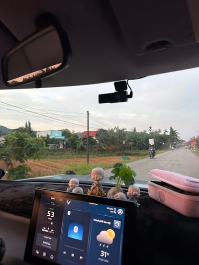 The image, rotated clockwise, provides an interior view from a car looking out onto a road in Bình Chương, Vietnam. On the left, a dashboard camera is mounted on the windshield, close to the rearview mirror. The dashboard is covered with a dark mat and adorned with three small monk figurines and a small potted plant. A pink pouch rests on the dashboard to the right.
Outside the car, a paved road extends into the distance, flanked by dense greenery, including palm trees, and what appears to be an agricultural field, possibly rice paddies. Power lines crisscross the sky, which is overcast, consistent with the weather information displayed. A motorcyclist is visible on the road ahead.
A car infotainment screen is prominent on the right, displaying digital information. The current time is "01:19". The car's status is "READY", with a speed of "53 km/h" and an odometer reading of "27179 km". The screen also shows the location as "Thành phố Tam Kỳ" (Tam Kỳ City), with a temperature of "31°C" and "mưa vừa" (light rain) indicated by a cloud and rain icon. A VinFast "V" logo is visible. Bluetooth is active, and a music player interface is shown, along with a battery status of "110-49%".