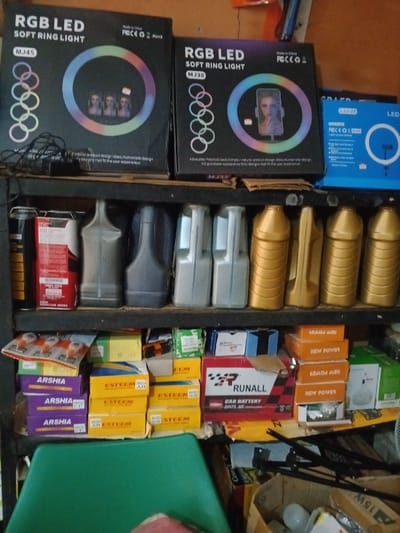 The image depicts the interior of a retail or supply establishment in Akodo, Nigeria. The scene is static, displaying various products on multiple shelves of a metal racking unit, suggesting a commercial setting dealing with electronics and automotive supplies.

On the top shelf, three boxed RGB LED soft ring lights are visible. The left box, labeled "MJ45," is black with an image of three female faces. The center box, labeled "MJ38," is also black and shows a smartphone held by a ring light, displaying a face. The right box is blue and labeled "LJ-D26."

The second shelf holds a row of plastic containers. From left to right: one black cylindrical item, one red and white rectangular container, followed by four identical grey plastic bottles with handles, and then five identical gold-colored plastic bottles, also with handles, likely containing automotive fluids such as motor oil.

The third shelf features stacked cardboard boxes and merchandise. Prominently, a red and black box labeled "RUNALL CAR BATTERY" is centrally located. To its left, several yellow boxes labeled "ESTEEM" and purple boxes labeled "ARSHIA" are stacked. Flat white packages containing multiple small, round objects, possibly light bulbs or components, are also visible. To the right of the RUNALL box, multiple orange boxes labeled "NEW POWER" are stacked.

The bottom portion of the image shows a partial view of a green plastic chair. Below the shelving, a cardboard box contains a white incandescent light bulb and other miscellaneous items. Dark metal rods are visible on the lower right, leaning against an unseen surface. The overall environment appears to be a storage or display area for a variety of consumer and automotive goods.