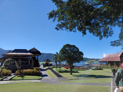 In Baturiti, Indonesia, a serene temple complex sits beside a tranquil lake under a clear blue sky. The scene is dominated by lush green grass, meticulously manicured flower beds bursting with pink and red blooms, and mature trees casting gentle shadows. Traditional Balinese architecture is evident in the structures with their distinctive thatched roofs and intricate carvings.
To the left, a temple building, adorned with decorative elements and surrounded by flowering plants, stands prominently. In the mid-ground, a path winds through the manicured lawns, leading towards a lake dotted with small boats. A large, full-bodied tree stands near the center of the frame, its branches reaching towards the sky.
On the right side, another building with a reddish-brown roof and open wooden pillars provides a shelter-like structure. A few individuals are visible. One person, seen from the back with shoulder-length red hair and wearing a green jacket, walks away from the camera. Further into the scene, another person in dark clothing walks along a pathway. The background features rolling, tree-covered hills that frame the shimmering blue lake. The overall atmosphere is one of peace and natural beauty.