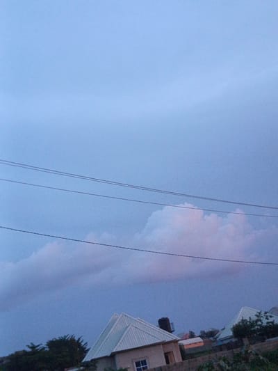 In the city of Rafin Bauna, Nigeria, an outdoor scene unfolds under a dusky, pale blue sky with wispy pink-tinged clouds. Several power lines stretch horizontally across the upper portion of the frame. In the lower half, the rooftops of several houses are visible. One house, on the left, has a white, gabled roof and a water tank. Another house, to the right, also features a white, gabled roof and is partially obscured by trees. The scene appears to be at dusk or dawn, with the sky transitioning between day and night.