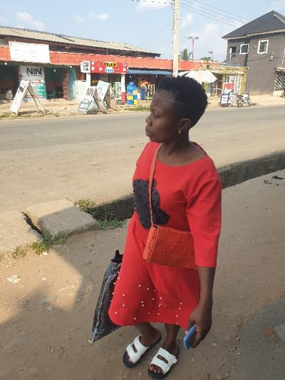 The image depicts a daytime street scene in Umuode, Nigeria. In the immediate foreground, a Black woman stands on a dirt and gravel roadside, facing right, slightly turned from the camera. She wears a red, mid-length dress with black accents on the chest and small white embellishments on the skirt, paired with white sandals with black soles. She carries an orange woven crossbody bag over her left shoulder and holds a blue smartphone in her right hand. A dark, possibly black, bag rests on the ground near her left leg.
Behind the woman, a paved asphalt road extends horizontally across the frame. On the far side of the road, to the left, a row of single-story commercial buildings with red corrugated metal roofs is visible. Prominent signage includes "NIN" and "GLO!", along with several A-frame signs. Yellow and blue plastic crates are stacked outside one establishment. To the right, further down the road, a dark-colored two-story building with white windows stands, followed by another commercial building with a white umbrella outside and a motorcycle parked nearby. The sky above is clear blue with minimal clouds. A utility pole and overhead wires are present in the background.