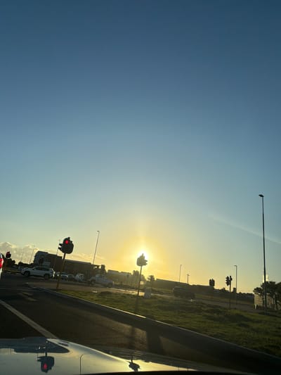 The image captures a street scene, likely taken from within a vehicle, during a sunrise or sunset, given the sun's position. The sky transitions from deep blue to a bright, golden-yellow hue near the horizon, where the sun shines intensely. The road stretches into the distance, bordered by a grassy area and buildings on one side. Several streetlights and traffic signals are visible along the roadside, their red lights indicating a stop. Cars are parked or lined up, suggesting an urban setting. The reflection of the traffic signal is captured by the car's window. I do not have enough information to determine the specific location in Cape Town, South Africa.