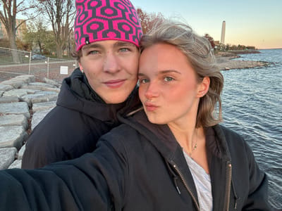 A selfie captures two individuals, a male and a female, standing beside a rocky shoreline in Kingston, Canada, with Lake Ontario in the background. The male, positioned on the left, wears a pink and black geometric-patterned beanie and a black puffer jacket, looking directly at the camera with a neutral expression. The female, slightly forward on the right, has light hair and a nose ring, wearing a dark zipped jacket over a light t-shirt and a beaded necklace; she poses with puckered lips towards the camera. The immediate environment includes a low formation of large, irregular grey rocks acting as a retaining wall or breakwater. In the mid-ground to the left, a metal fence separates a grassy area with sparse trees from the shoreline. The body of water, Lake Ontario, extends across the right and far background, reflecting the late afternoon light from a pale blue sky. A prominent, slender white chimney, identifiable as belonging to the Kingston General Hospital power plant, stands tall in the distant background to the right, over the water.