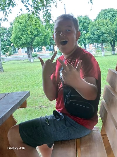 A young boy with light skin and short, dark hair is sitting on a wooden bench outdoors. He is wearing a red t-shirt and dark shorts. A black fanny pack is worn across his chest. He is making a hand gesture with both hands, extending his index and middle fingers. The boy is looking towards the camera with his mouth open. He is positioned in front of a grassy area with trees and a building in the background. The scene appears to be in Ustyvytsia, Ukraine.