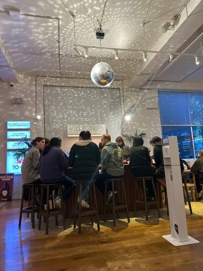This image depicts the interior of a Yo-Chi frozen yogurt establishment located in Melbourne, Australia. The central element of the scene is a dark wooden bar counter extending across the mid-ground, where six individuals are seated on tall wooden stools.

From left to right, the seated individuals include: a female with light-brown hair wearing a grey top, seated in profile and conversing with a female to her right who has dark hair and wears a dark purple top and blue jeans, with her back mostly to the camera. Next is an individual with their back to the camera, wearing a dark puffer jacket and light blue jeans. Adjacent is a male with short light-brown hair, wearing a light green hooded sweatshirt with a graphic design on the back. Further right, an individual with curly light-brown hair wears a dark jacket, and another individual in a dark jacket is partially visible. These individuals appear to be engaged in conversation or awaiting service.

The ceiling is white, fitted with exposed conduits and multiple white track lights. A large disco ball hangs centrally from the ceiling, illuminated by an overhead projector, casting numerous small, circular light reflections across the white walls and ceiling. A large window with horizontal blinds occupies the background, partially revealing faint exterior light. Another window on the right side also shows exterior lighting. The flooring consists of polished, light-toned wooden planks, reflecting ambient light.

On the left wall, two illuminated signs are visible. The upper sign reads, "REAL YOGURT WITH PROBIOTICS AÇAI FROM ORGANIC BERRIES," and below it, "UNIQUE TOPPINGS FRESH FRUIT CUT DAILY." A lower sign partially visible states "NEW AÇAÍ / YO-CHI". A tall, white, freestanding hand sanitiser dispenser, branded with "YO-CHI", is positioned on the right side of the foreground. A potted plant is situated behind the bar counter.
