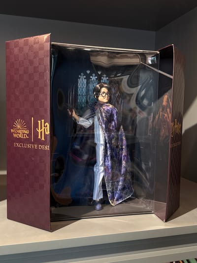 This media file features a Harry Potter doll, dressed in a blue and white striped shirt and a shimmering dark purple cloak with gold accents, lying within a clear plastic display box. The doll has dark hair and wears round glasses. The box itself is designed to resemble a book, with a checkered burgundy pattern and gold lettering on the exterior. Visible text on the box includes "Harry Potter," "GN COLLECTION," "WIZARDING WORLD," and "EXCLUSIVE DESI." The doll appears to be posed as if holding a wand. The setting is a dimly lit indoor environment, possibly a store or a personal collection display. The lighting suggests it may be daytime, with light casting shadows on the box and its contents. No specific location cues are visible beyond the implied context of a Harry Potter merchandise display. The overall impression is of a collectible item presented for viewing or sale.