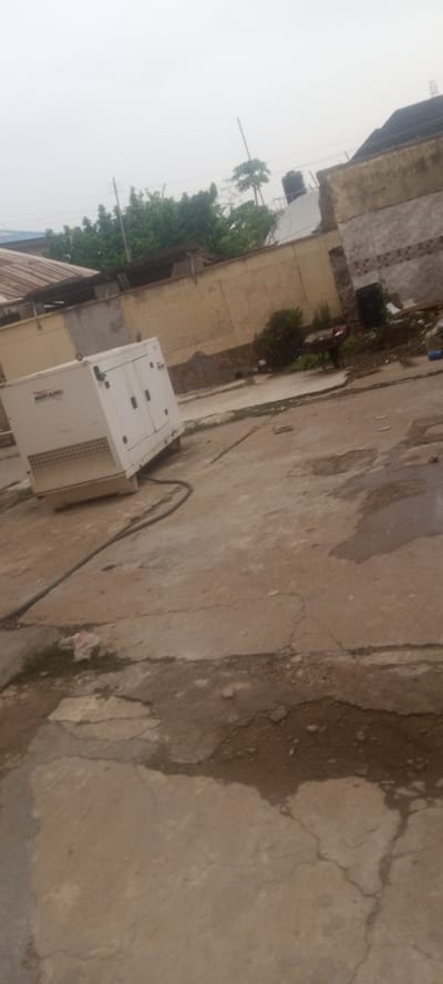 The scene shows an outdoor area in what appears to be Kaduna, Nigeria. A white, boxed generator labeled "MIKANO" sits on cracked concrete. A thick black cable runs from the generator. Behind the generator is a beige wall. Debris and overgrown foliage are present near the wall. The sky is overcast. The buildings in the background seem old.