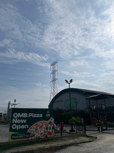 The image depicts an outdoor scene in Ilado, Nigeria, characterized by a bright, cloudy sky. Dominating the background is a tall, lattice-like electricity pylon with multiple transmission lines stretching across the frame. To the right, a modern, curvilinear building with a dark green roof and grey walls is visible, featuring a prominent circular green motif on its facade.
In the foreground, a large, dark green billboard announces "QMB Pizza Now Open". The advertisement includes an appetizing image of a pizza, the "QMB restaurant" logo, and a contact number: "Call/WhatsApp 0806 923 7750". Adjacent to the billboard, a paved area with some greenery and a low wall suggests an outdoor seating or entrance area for the restaurant. Scattered streetlights and fences are also present in the scene. The overall impression is a commercial area with a focus on promoting a new pizza establishment.