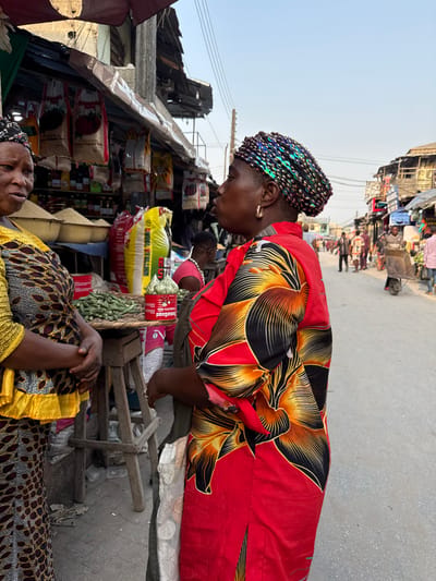 Two women at an outdoor market. One in a red floral dress and iridescent headdress leans over a vendor stall stacked with food supplies. Another woman in a yellow and brown patterned dress is reclining, eyes closed, at the bottom of the frame. Various market stalls and people are visible in the background, under a clear sky. The image is taken from a slightly elevated angle.