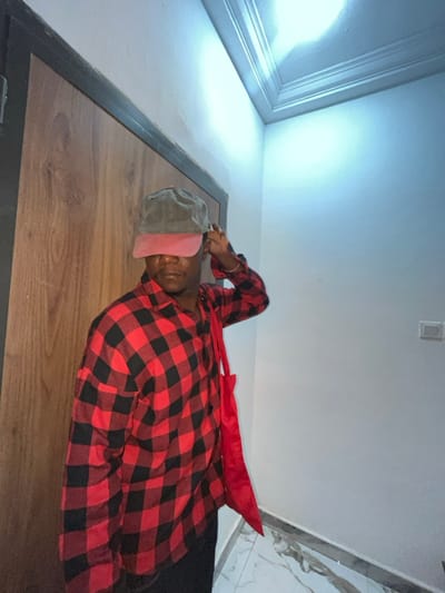 A man lies on a wooden floor, clad in a red and black checkered shirt. He wears a cap with a pink visor covering his eyes. A long red scarf lies across his shoulder. The camera's low angle shows the ceiling, which features a light fixture and decorative molding. A wall with marble tile is visible in the background. The room is dimly lit.