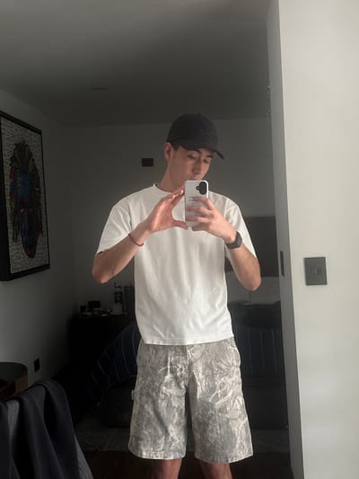 A person wearing a white t-shirt, camouflage shorts, and a black baseball cap is taking a mirror selfie in what appears to be a bedroom. The person is holding a white smartphone in their right hand, capturing their reflection. On their left wrist, they wear a black fitness tracker, and on their right wrist, a thin red bracelet. The background includes a framed artwork on the wall to the left, a dark dresser with items on it, and a bed with striped bedding. To the right, a light-colored wall is visible, with a dark light switch plate on it. The overall lighting suggests an indoor setting during the day, with soft light. There is no discernible evidence linking this image to San Bernardino Tlaxcalancingo, Mexico; it is a general interior scene.