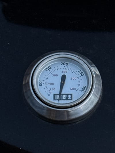 The image shows a close-up of a Weber thermometer on a dark, possibly black, surface. The thermometer has a circular dial with markings in both Fahrenheit and Celsius. The needle is pointing just above the 400°F mark, indicating a temperature of approximately 400-420°F. The Weber logo, featuring the brand name and a grill icon, is visible on the bottom part of the dial. The thermometer appears to be built into a grill or other cooking apparatus. There is no indication of the city of Bruges, Belgium, in the image or its context.
