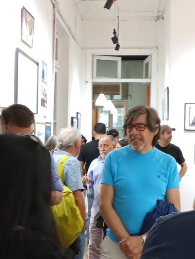 A crowd gathers in an art gallery. A man in a bright blue shirt and glasses stands towards the front, smiling. Behind him, another man holds a champagne flute. A light yellow backpack is visible on a person in a light blue shirt. Several people are positioned in the background with a white doorway in the distance. Pictures hang on the white walls. Overhead, black lights are suspended.