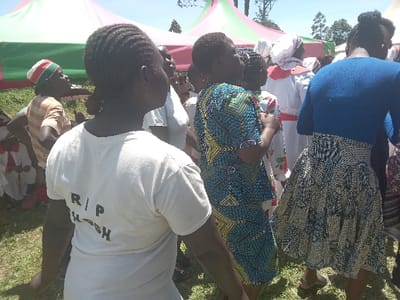 The image shows a gathering of people in Malava, Kenya (latitude: 0.515, longitude: 34.827). The scene is outside and features a group of individuals, some wearing traditional clothing, standing near a white tent. The central figure, a young woman with braided hair, is wearing a white t-shirt with "RIP WASH" printed on the back. She is surrounded by other women and a few men. One woman is wearing a white dress with red trim and another woman wears a blue dress with a patterned skirt. The grass in the background is green and the sky is clear and blue.  The event appears to be a somber occasion, potentially a funeral or memorial service.