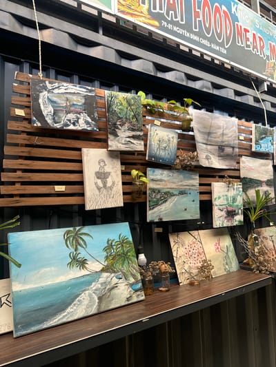 In Phan Thiết, Vietnam, this image depicts an outdoor art display on a wooden slatted wall, likely attached to shipping containers as evidenced by the dark, corrugated background. The display features various paintings and sketches. In the foreground, a painting of a tropical beach with palm trees and crashing waves is prominently displayed on a dark wooden surface. Above this, a series of paintings are hung on the wooden slats. These include a pencil sketch of a person carrying a basket, a landscape with a body of water and sailboats, a forest scene with a waterfall, and abstract pieces with floral motifs. Several small yellow price tags are visible. Small jars with plants and dried flowers are placed among the artwork, adding a decorative touch. A sign with Vietnamese text and English phrases like "THAI FOOD" and "KING OF SANDWICH" is visible in the background, indicating the presence of a food establishment nearby. The overall setting suggests an art market or an outdoor gallery attached to a commercial establishment in a coastal area.
