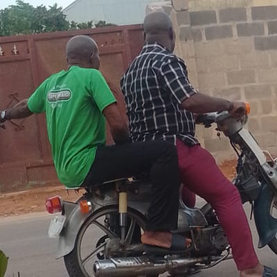 In Tempure, Nigeria, two men are riding on a motorcycle. The man in front is wearing a bright green t-shirt with a logo that says "CEM RAID". He has dark skin and a shaved head. The man behind him is wearing a black and white checkered shirt and dark pants. He has dark skin and a shaved head. They are on a paved road, with a brown fence and brick wall in the background. The motorcycle appears to be an older model. The lighting suggests it is daytime.