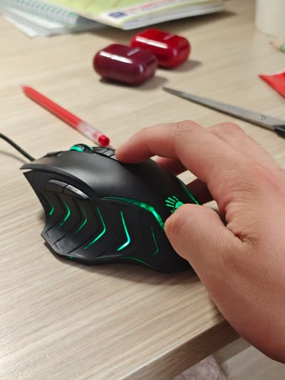 A close-up shot shows a person's hand operating a black gaming mouse with green LED accents. The mouse is positioned on a light-colored wooden desk. In the background, out of focus, are a red pen, two red objects that appear to be containers or caps, a stack of notebooks, and a pair of scissors. The scene suggests a workspace or home office setting, possibly in Svietlahorsk, Belarus. The lighting is artificial and illuminates the desk surface and the mouse's glowing features. No specific time of day or weather conditions are discernible from the image. The focus is entirely on the interaction between the hand and the mouse, implying an activity such as gaming or computer work. No discernible text is visible in the image.