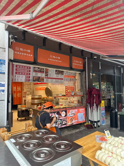 The image depicts a street food stall in the city of Chongqing High-tech Zone, China, under a red and white striped awning. The stall specializes in steamed buns and other food items, as indicated by the Chinese signage and the display of food products. A person wearing an orange cap and apron is seen working behind the counter, preparing food. In the foreground, several stainless steel steaming pots are visible, along with stacks of packaged steamed buns on a wooden table. The background shows a shopfront with more signage and a glass door. The overall scene is that of a bustling food vendor operating in an urban environment.