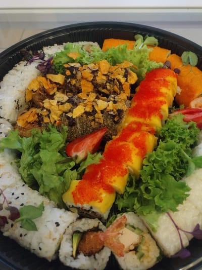 In Crauthem, Luxembourg, a black plastic tray holds a platter of sushi. The tray is filled with various types of sushi rolls, artfully arranged with fresh green lettuce. A prominent sushi roll in the center is covered in bright orange tobiko (flying fish roe) and filled with what appears to be mango and possibly avocado. To the left, another section of the platter features sushi topped with crispy fried onion flakes. Some sushi rolls on the left and right sides are decorated with black and white sesame seeds. A halved strawberry is placed near the center of the platter, adding a touch of red color. Microgreens and thin purple sprouts are scattered as garnishes. The overall presentation suggests a prepared meal, likely for takeout or catering, given the disposable nature of the tray. The lighting indicates an indoor setting, possibly a kitchen or dining area. There are no people visible in the image.
