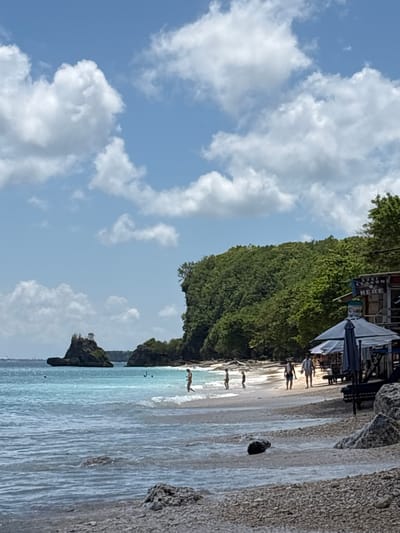 The image captures a beach scene in Uluwatu, Indonesia. The frame is vertically oriented.
A lush green cliff face descends towards the beach, topped with dense vegetation. The beach is light tan, and the water transitions from turquoise to a deeper blue further out. Several people are visible along the shoreline; some are walking, and a few appear to be swimming in the water. The sky above is blue with scattered white clouds. Along the top edge, there appears to be a small structure with umbrellas, potentially a beachside cafe or shop. "Surf lesson here" is partially visible. A rocky outcropping is visible on the right side of the frame. The image suggests a tropical, coastal environment with leisure activities.