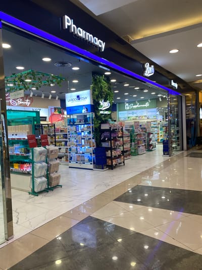 A retail store interior, likely a pharmacy or a beauty and health store, is depicted in this image. The storefront signage prominently displays "Pharmacy" in large white letters, with a blue illuminated strip below. To the right, a sign for "Boots" is also visible. The store is well-lit with overhead lights. Inside, shelves are stocked with various products, including skincare items, supplements, and other health and beauty products. A section of the store appears to be dedicated to "La Roche-Posay" products. Decorative elements like artificial plants and greenery are incorporated into the store's design. The flooring is a marble-patterned tile with geometric divisions of light and dark colors. The overall impression is of a clean, organized, and modern retail environment. The location is West Jakarta, Indonesia.