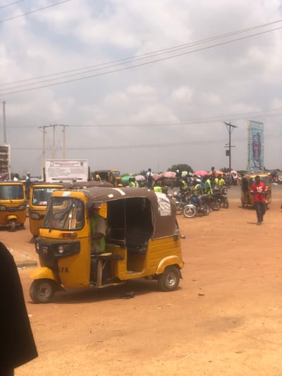 The scene depicts an outdoor transportation and commercial area in Lafia, Nigeria. The ground is unpaved, consisting of reddish-brown soil. The sky is partly cloudy with visible power lines and utility poles.
In the left foreground, a yellow three-wheeled motorized rickshaw (keke napep) with a brown fabric canopy is stationary, facing forward and slightly left. The letters "LFA" are visible on its front fender. A male driver, wearing a light-colored shirt and a green safety vest, is seated inside. A small, unadorned wooden tray is affixed to the exterior of the vehicle's left side. To the right of the foreground rickshaw, a male individual wearing a red shirt with the number "15" emblazoned on the front walks from right to left across the mid-ground.
In the mid-ground and background, multiple additional yellow three-wheeled rickshaws are parked. To the right, a cluster of motorcycles is stationary, accompanied by several individuals, many of whom are wearing bright green safety vests. Colorful market umbrellas (pink, green, blue) are visible in the background, indicating market stalls or shaded gathering points. A white rectangular sign board with unreadable text is mounted on poles in the left-center background. On the far right, a tall, blue-themed billboard displays a stylized image of a person's head. Numerous other people are dispersed throughout the background, appearing to be engaged in various activities, consistent with a transport hub or market.