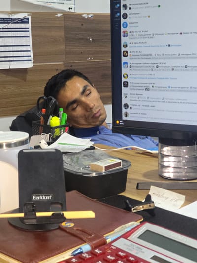 The image shows a man sleeping at a desk in an office. The man is lying down with his head resting on his arms, which are on the desk. There are various office supplies on the desk, including pens, scissors, a stapler, a calculator, and a mobile phone stand. A computer monitor is visible in the background, displaying a variety of icons and text, some of which appear to be in Cyrillic script. The office environment appears to be somewhat cluttered. The image does not provide any specific clues about the city of Karshi, Uzbekistan, but the presence of Cyrillic script on the monitor suggests a location within a region where this script is used, which could include Uzbekistan. The time of day appears to be daytime, indicated by the lighting.