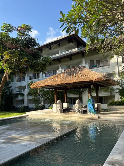 The image depicts a building in Jimbaran, Indonesia. The structure is multi-storied and white, with dark accents. A thatched-roof shelter stands adjacent to the building, supported by wooden columns. The shelter appears to house stacked chairs. To the right of the shelter is a decorative water feature with a textured surface. Lush greenery surrounds the building, including trees with visible red flowers. A clear blue sky is visible above, partly obscured by the foliage. Sculptures of animals are present on the right side of the shelter.
